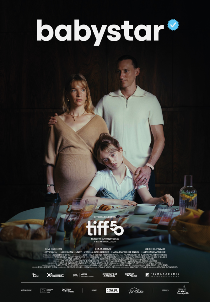 Plakat utrzymany w ciemnej, stonowanej kolorystyce. Na tle ciemnej, drewnianej ściany stoją trzy osoby: kobieta w ciąży, mężczyzna oraz młoda dziewczyna. Kobieta (blond włosy, beżowa sukienka) stoi po lewej i patrzy przed siebie z poważnym wyrazem twarzy. Mężczyzna (krótkie włosy, jasna koszulka polo) stoi obok niej, trzyma rękę na jej ramieniu i patrzy w jej stronę. Przed nimi, przy stole, siedzi dziewczyna (nastolatka), oparta głową o rękę, z wyraźnie smutnym lub znużonym wyrazem twarzy. Na stole znajdują się resztki śniadania: talerze, szklanki z napojami, dzbanek oraz jedzenie. Na górze widnieje duży napis „babystar” z symbolem weryfikacji. Na dole znajdują się informacje o festiwalu (TIFF) oraz twórcach filmu. Plakat sugeruje napiętą, emocjonalną relację rodzinną.